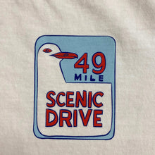 Load image into Gallery viewer, A close-up of a white tee shirt surface featuring a printed graphic with a white seagull, red text reading '49 MILE SCENIC DRIVE,' and a light blue background inside the design.